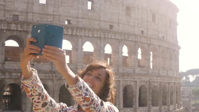 Beautiful young woman in colorful fashion dress alone on hill with bike taking selfies with colosseum in Rome with smartphone at sunset attractive elegant girl