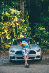 Young man standing by the Ford Mustang GT on the island of Kauai, Hawaii holding a coconut in his...