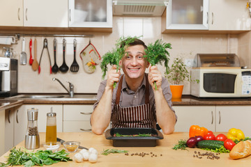 Handsome fun young man in apron sitting at table with dill or fennel, cooking at home preparing meat stake from pork, beef or lamb, in light kitchen with wooden surface, full of fancy kitchenware.