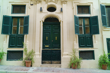 Fototapeta na wymiar Traditional classic style wooden medieval vintage facade and green painted doors with knockers and windows in Valletta, Malta. Green plants in pots near stone wall.