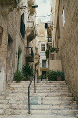 Medieval town narrow street with stairs in the city Valletta on the island of Malta. Yellowbalconies and doors and green plants near traditional  stone buildings. Analog camera toned image.