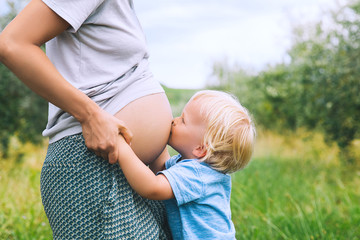 Child boy kissing belly of pregnant her mother on nature background.