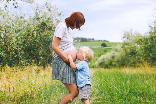 Child boy kissing belly of pregnant her mother on nature background.