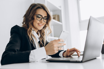 Young attractive business woman working in her office with a laptop, looks into the phone and smiling