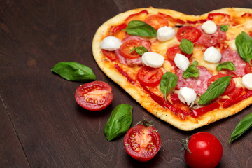 Tomatoes and Pizza in shape heart with salami, , basil and mozzarella against a dark wooden brown background.Copy space.Top View. Creative valentine day background.Love concept.Closeup.Selective focus