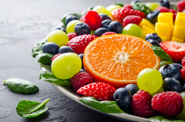 Fruit platter with various fresh strawberry, raspberry, blueberry, tangerine, grape, mango, spinach on a dark black stone background. Copy space, front view, horizontal image