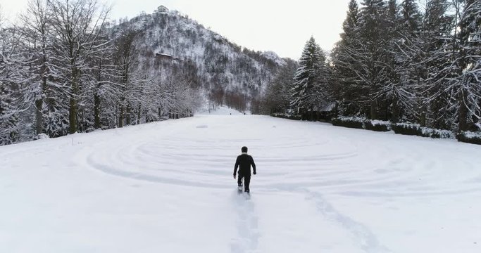 forward aerial over man walking with snowshoes on mountain snow covered field near pine forest woods in overcast winter.Europe Alps outdoor nature scape snowy mountains.Back following.4k drone flight