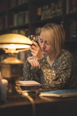 Mature blonde lady writing down notes, studying or writing a book and holding a glass of wine in an intimate private library surrounding