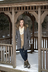 Beautiful brunette young woman in winter scene - snow covered park wearing winter clothes - layered sweaters and parka standing in gazebo or shelter