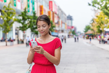 Asian woman outside on Wangfujing shopping street in Beijing, china, using mobile phone app to shop online. Happy Chinese woman texting sms on smartphone.