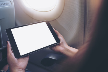 Mockup image of a woman holding and looking at black tablet pc with blank white desktop screen next to an airplane window with clouds and sky background
