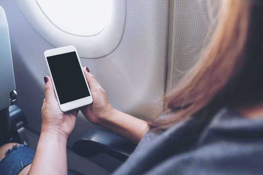 Mockup image of a woman holding and looking at white smart phone with blank black desktop screen next to an airplane window with clouds and sky background