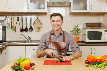 Handsome smiling caucasian young man in an apron, brown shirt sitting at table, cuts vegetable for salad with knife in light kitchen. Dieting concept. Healthy lifestyle. Cooking at home. Prepare food.