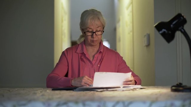 A bewildered female senior citizen on a fixed income sitting at a table who opens her check book to start paying a bill she is concerned about.