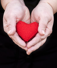 couple of hands of elderly man with wrinkled hands gently holding knitted red heart on a black background