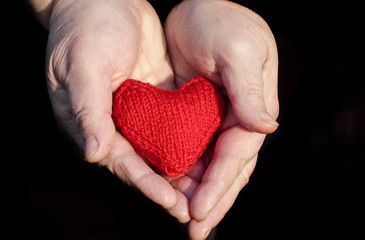 Obraz premium couple of hands of an elderly pensioner with wrinkled hands gently holding knitted red heart on a black background