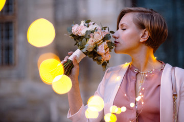 Beautiful woman inhaling fragrance of a flower. Evening portrait with glimmer garland bokeh. Close...