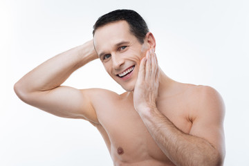 Obraz premium Feeling positive. Joyful cheerful nice man smiling and showing his emotions while touching his cheek