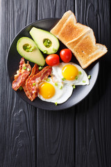 Delicious breakfast of eggs with crispy bacon, avocado, toast and tomatoes close-up. Vertical top view