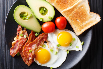hearty breakfast: fried eggs with bacon, avocado, toast and tomatoes close-up on a plate. horizontal top view