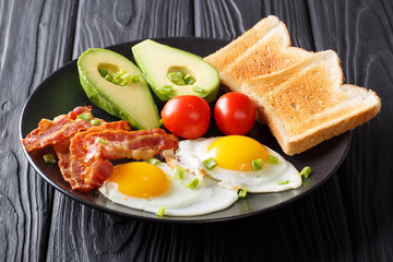 Freshly fried eggs with crispy bacon, avocado, toast and tomatoes close-up on a black plate on a table. horizontal
