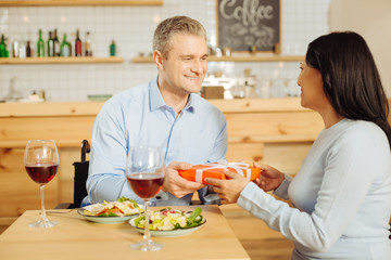For you. Handsome content handicapped man smiling and giving a present to his attractive beloved alert woman while having romantic dinner