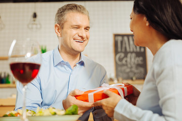 Gift for you. Attractive inspired well-built man smiling and giving a present to his beautiful beloved happy woman while having romantic dinner