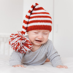 portrait of little crying infant child in funny knitted hat lying on bed