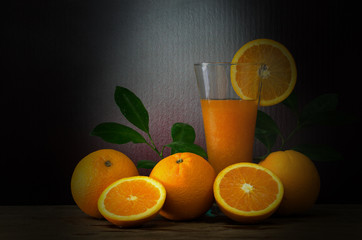 Fresh oranges with green leaf and juice in glass on wooden plate in dark room wich has light splash down to pile of oranges