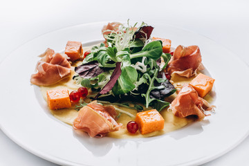 Salad of jamon, spinach, arugula, basil, cress salad and sauce, with grapefruit grains, on a round white plate. Side view