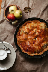 Traditional sweet apple pie on red wooden background with small bowl of cream on a piece of linen fabric. Harvest baking concept.