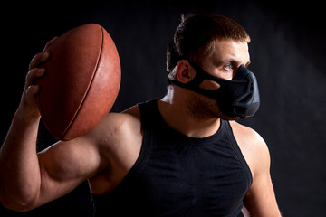A dark-haired male athlete in a black training mask, a sports shirt holding a rugby ball and waving it on a black isolated background
