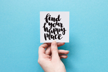 Minimal composition on a blue background with girl's hand holding card with quote "find your happy place" written in calligraphy style