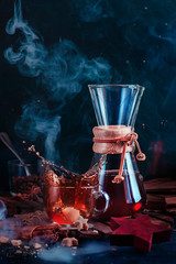 Glass coffee cup with a dynamic splash and rising steam. Hourglass-shaped coffeemaker on a dark background with copy space. Action food photography. Alternative coffee brewing concept.