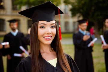 Happy cute chinese brunette girl is smiling, blurred class mates with diplomas behind. She is in a...