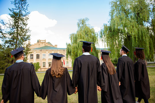 Rear view of six successful international young graduates in black robes and head wear, finished their education, holding hands and bonding, looking at the university building, nice sunny summer day