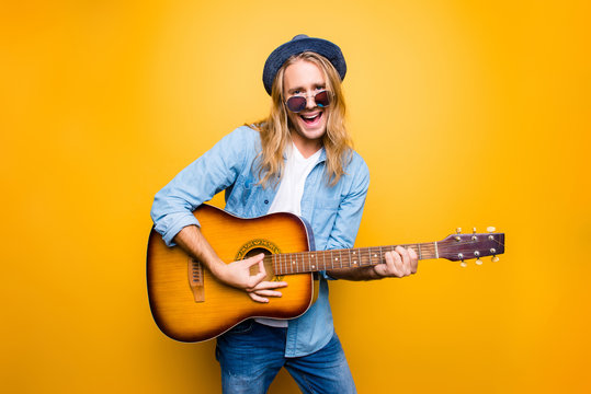 Portrait of active attractive young guitarist man playing on guitar singing his favourite song dancing like crazy over yellow background
