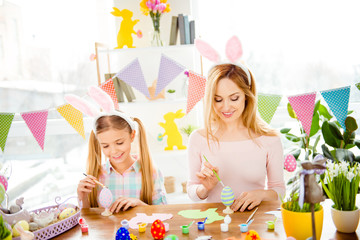 Happy Easter atmosphere! Funny, pretty mummy teaching, training her cute, joyful, small daughter draw, paint, handmade decorate easter eggs, together wearing bunny ears