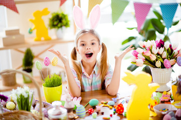 Cute, pretty, impressed, little child with wide open mouth, gesturing with hands, celebrating Easter, wearing bunny ears, costume, sitting at decorated desk