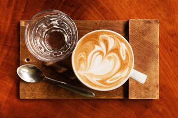 Cup of hot cappuccino and glass of water on the wooden table in a cafe, top view.