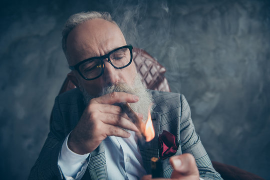 Close up portrait of attractive, ideal, stunning, brutal, harsh, old man, dandy in glasses, tux setting on fire cigar over gray background