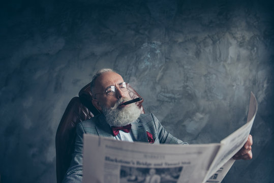 Portrait of brutal attractive old man in tuxedo holding, looking at newspaper, reading news about finance, politic, economy, smoking cigar, sitting in armchair, gray background