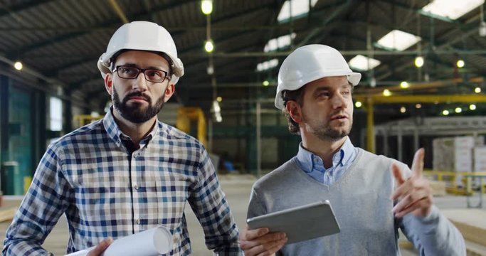 Two Caucasian men in helmets walking in the big factory room, talking and looking at the tablet screen, having a discussion. Indoors. Portrait. Close up