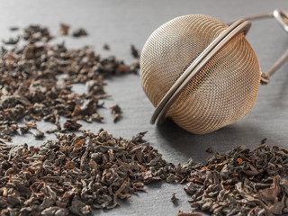 Round metal tea strainer for brewing tea and the dry leaves of black tea on a black background