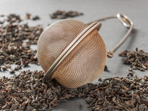 Round metal tea strainer for brewing tea and the dry leaves of black tea on a black background