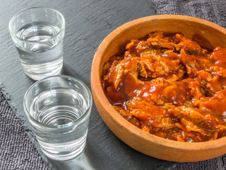 Sprat in tomato sauce in a ceramic dish and two glasses of vodka on a black background. Canned fish.