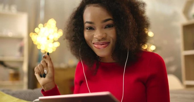 Attractive young African woman in red sweater sitting on sofa and listening music in headphones on background of beautiful New Year’s tree with lights. Enjoying. Evening.