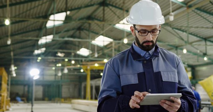 Caucasian male factory worker walking with a tablet device in his hands. Manufactory space. Indoors. Portrait