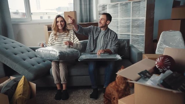 Young people moving to new apartment. Nice couple in love unpacking purchases. Lot of sealed boxes in room. Couple talking about where to put present picture.