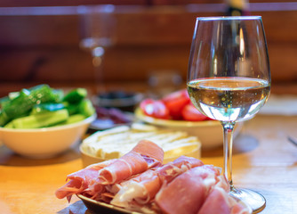 Sliced prosciutto with a glass of wine and vegetables in the background.